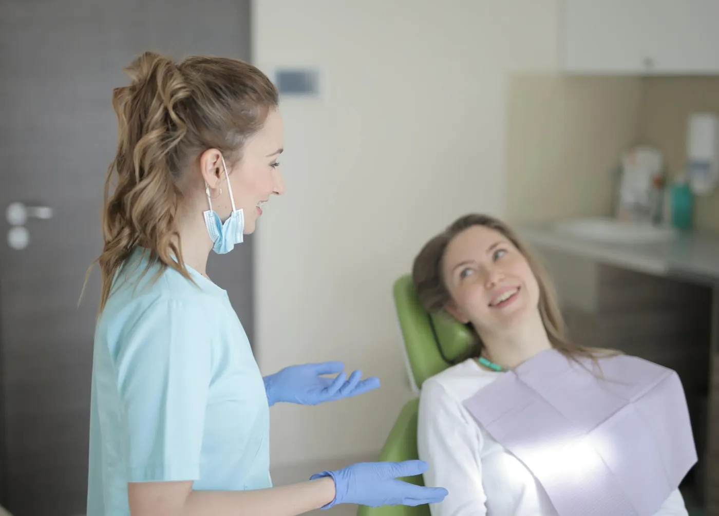 Positive young female dentist in latex gloves and uniform talking with young patient while sitting near chair and preparing for treatment in modern dental room in hospital